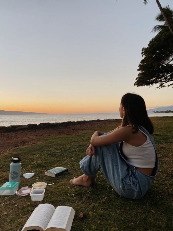 A girl sitting in a field watching as the sun sets in the horizon